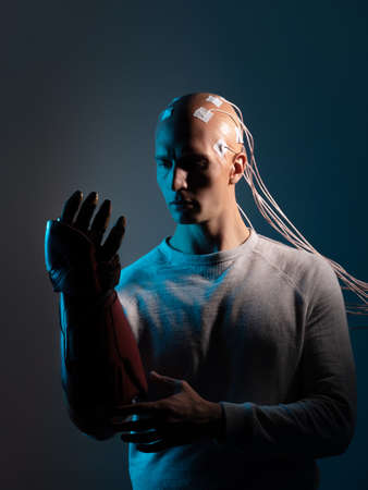 young man with a head entangled with wires and with a robotic arm, augmentations of the human body. contacts on the bald, signals to the brain. Neurointerface, future technologies, creative concept.の写真素材
