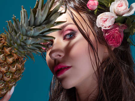 A beautiful young brunette with bright blue eyes, in a Hawaiian image, with pineapple in hands. Wet hair and flowers in the hairstyle. Mysterious portrait in the shadowsの写真素材