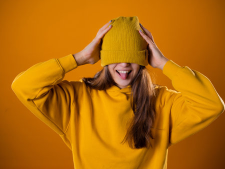 A young woman in a yellow hoodie and a hat on a yellow background. Cute brunette pulled a hat over her eyes, close-up portraitの写真素材