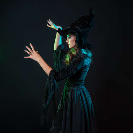 A witch creates magic, a young beautiful brunette in a pointed hat makes passes with her hands, spells. Halloween Costume Partyの写真素材