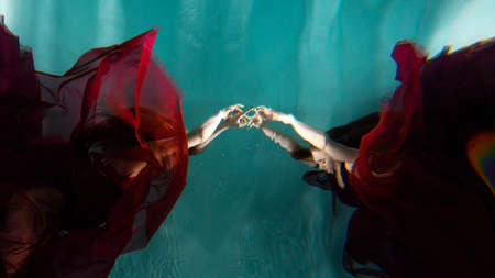 Photo underwater, a young beautiful woman in red with red hair reaches for the surface of the water, a human and his reflection. mystical underwater portraitの写真素材