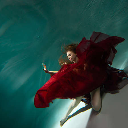 Beautiful underwater shooting, a young woman in a red dress with a long skirt swimming underwater, distortion and caustic effects.の写真素材