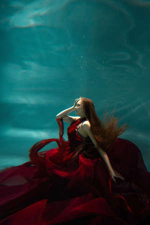 Beautiful underwater shooting, a young woman in a red dress with a long skirt swimming underwater, distortion and caustic effects.の写真素材