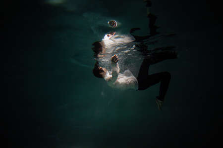 Sink. A young guy in a white shirt falls into the water, a photo from under the water. The concept of falling down, diving to the depth, contrasting dark photoの写真素材