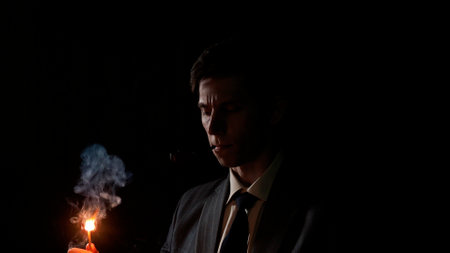 A serious young man lights his pipe with a match, a man in a gray suit with a tie holds a burning match in his hand, deep shadows in a retro noir styleの写真素材