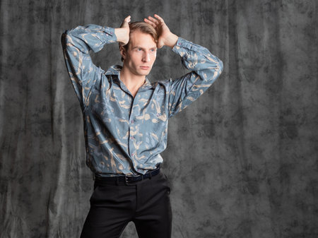 Stylish elegant young man in a blue silk shirt posing in the studio on a gray fabric backgroundの写真素材