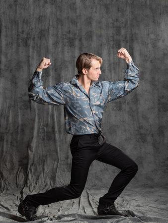 Stylish elegant young man in a blue silk shirt posing in the studio on a gray fabric background. brutal poseの写真素材