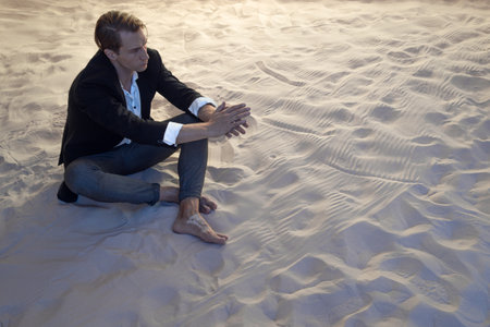 sad financier is frustrated, a young man with big plans is faced with a global economic crisis, concept. businessman in a suit is sitting on a pile of sandの写真素材
