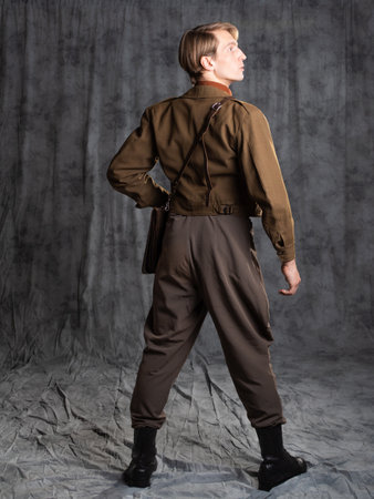 A young guy in military-style clothes, a brown flight jacket and breeches with suspenders. Posing in...