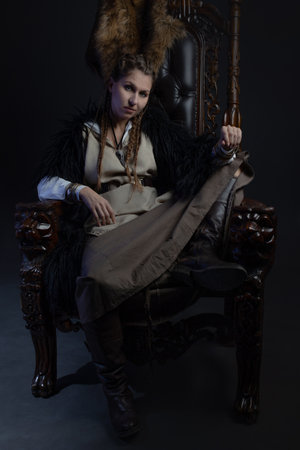An authoritative, self-confident woman in a Viking-style fantasy costume, sitting on a high chair or...