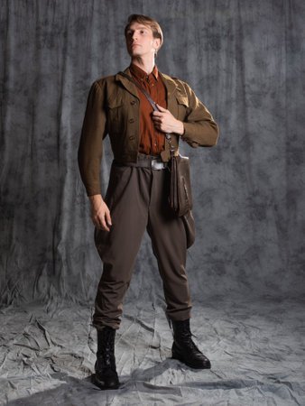 A young guy in military-style clothes, a brown flight jacket and breeches with suspenders. Posing in...