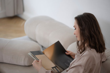 stylish and confident brunette is sitting on the couch and reading a magazine, a comfortable...