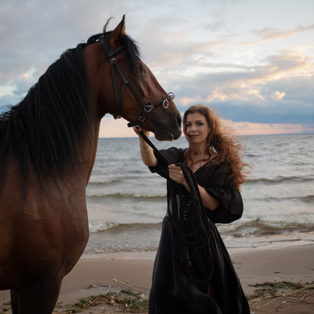 beautiful young rider in a dress and with her hair down, leads her horse along the beach. horseback riding in the open airの写真素材