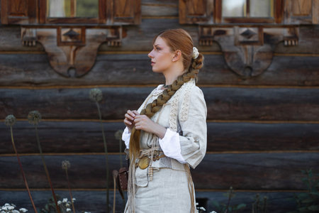 a young girl with a long blond braid in a folk costume, in a village among wooden architecture,...