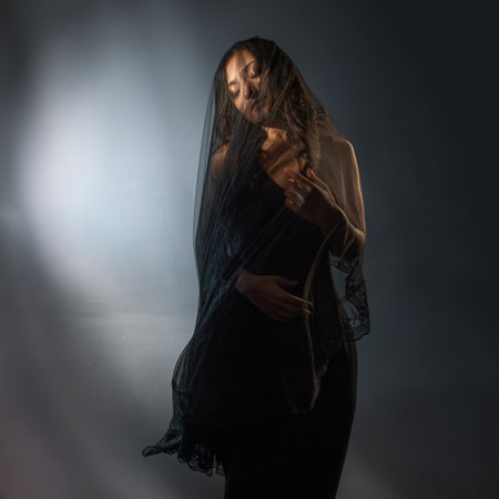 beautiful image in the Gothic style shows a young woman with black hair, wearing a black dress and a veil that hides her face. The folds of the veil drape around her figure in a mysterious way, creating a sense of mystery.の写真素材