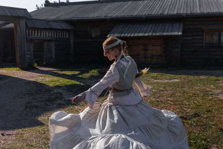 A young girl with a long braid in a folk costume is dancing and having fun in a village among wooden architecture, Slavic or Nordic styleの写真素材