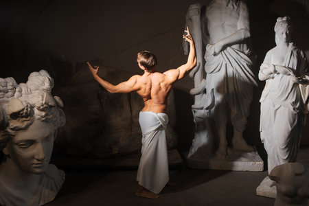 A sculptors workshop, a muscular man in a tunic among antique statues. Bodybuilder and model, aesthetics of harmonious development of the bodyの写真素材