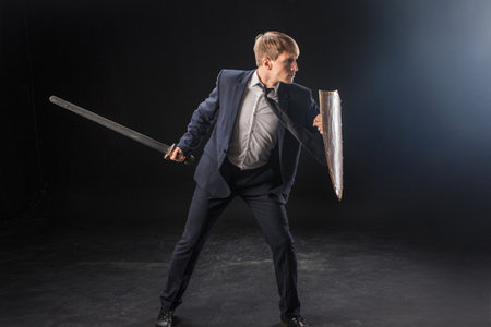 In a dramatic studio pose, a man in a navy suit stands poised with a sword and shield. The dark background emphasizes the contrast between corporate and medieval imagery.の写真素材