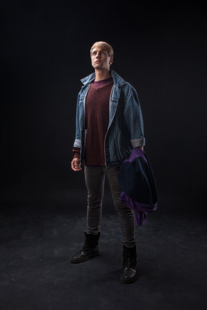 person stands confidently wearing a denim jacket over a maroon sweater, holding a blue jacket in a studio with a dark, atmospheric background. Casual and stylish look.の写真素材
