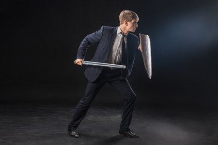 In a dramatic studio pose, a man in a navy suit stands poised with a sword and shield. The dark background emphasizes the contrast between corporate and medieval imagery.の写真素材
