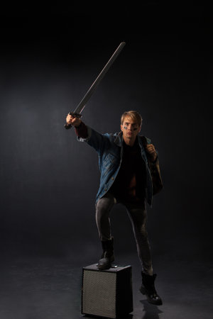 young student in a denim jacket and maroon sweater stands confidently in a studio, holding books and a sword, embodying a blend of geek and gamer culture.の写真素材