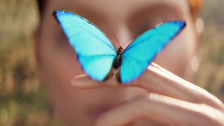 A young woman looks at a beautiful butterfly, a blue swallowtail, sitting on her finger, close-up. scene symbolizes unity with nature, showcasing vibrant greenery, soft sunlight filtering through trees, and a serene atmosphere.の写真素材