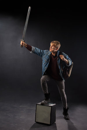 young student in a denim jacket and maroon sweater stands confidently in a studio, holding books and a sword, embodying a blend of geek and gamer culture.の写真素材