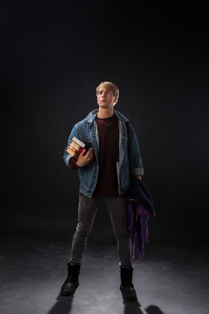 In a dimly lit studio, a person in a denim jacket and burgundy sweater raises a hand with intent. They hold books and a purple jacket, embodying youthful exploration.の写真素材