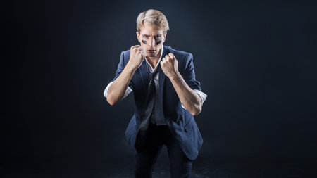 In a dimly lit studio, a man in a navy suit adopts a boxing stance, showcasing strength and determination. The sleeves are casually rolled up, enhancing his confident, dynamic pose.の写真素材