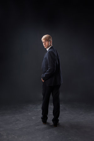 In a dark studio, a man stands confidently in a crisp black suit and tie. The dramatic lighting highlights his attire against the black backdrop, emphasizing formality and power.の写真素材