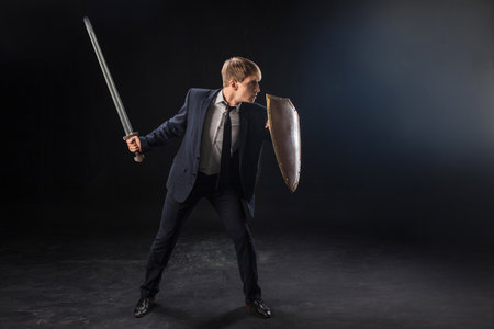 In a dramatic studio pose, a man in a navy suit stands poised with a sword and shield. The dark background emphasizes the contrast between corporate and medieval imagery.の写真素材
