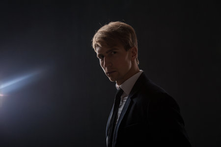 In a dark studio, a man in a black suit and white shirt stands in profile. The contrast between his attire and the shadowy background creates an air of mystery and professionalism.の写真素材