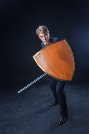 In a dramatic studio pose, a man in a navy suit stands poised with a sword and shield. The dark background emphasizes the contrast between corporate and medieval imagery.の写真素材