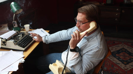 Male author at vintage desk talking on wired phone, typewriter and papers in cinematic soft-focus office scene.の写真素材