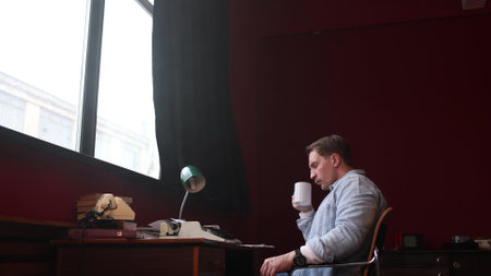 Male author experiences writers block at his wooden desk, surrounded by papers and books in softly...