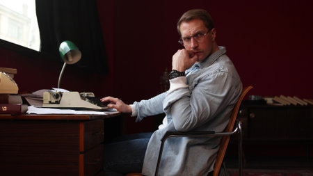 Writer deep in creative thought at his wooden desk, surrounded by books and papers in soft focus.の写真素材