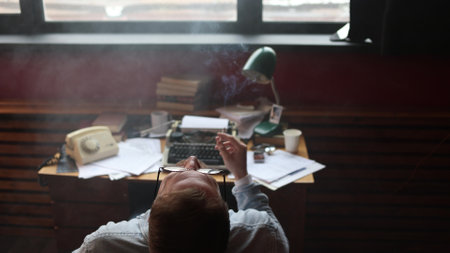 Young journalist smokes in frustration at vintage desk, cinematic soft lighting highlighting typewriter work.の写真素材