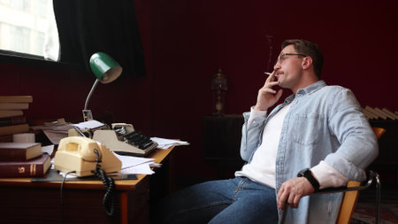 Young press journalist smokes while writing intensely, vintage office with cinematic soft-focus details.の写真素材