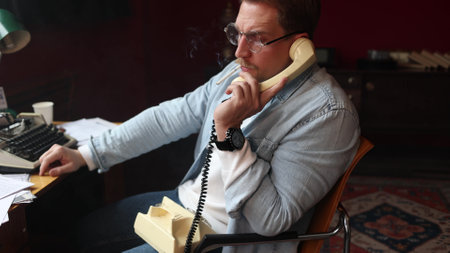 Male author at vintage desk, phone in hand with typewriter and papers in cinematic soft-focusの写真素材