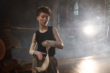 A woman in a boxing ring, wrapping an elastic bandage around her arms, preparing for boxing training. The photo is in a cinematic style, the light gives the mood to a sports motivational story about self-improvement and the path to the goalの写真素材