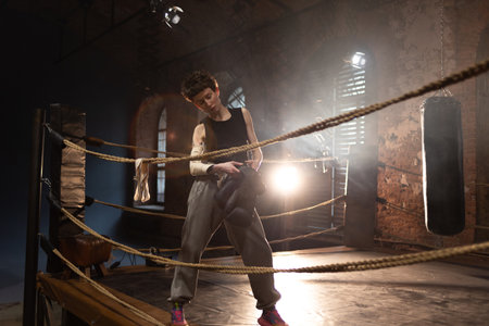 A woman in a boxing ring, holding boxing gloves in her hands, preparing for boxing training. The photo is in a cinematic style, the light gives the mood to a sports motivational story about self-improvement and the path to the goalの写真素材
