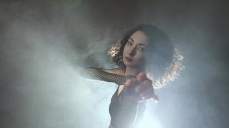Modern dance, a young woman dancing on a dark background, atmospheric lighting in smoke. A girl in black pants and a mesh blouse performs modern movements in heelsの写真素材