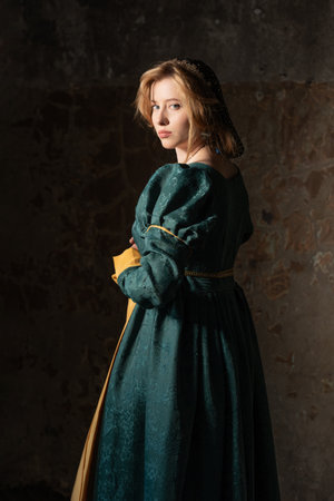 Elegant young woman in green medieval gown, golden hair flowing, standing before weathered stone backdrop.の写真素材