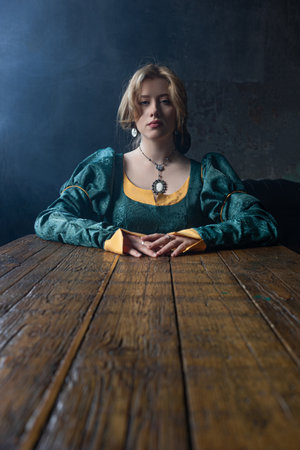 Elegant young lady in green medieval gown sits gracefully at rustic table, historical aestheticの写真素材