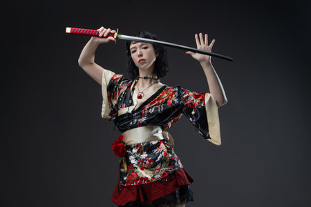 A mysterious brunette femme fatale poses in a short kimono blended with cyberpunk elements, holding a katana sword against a neutral gray studio backgroundの写真素材