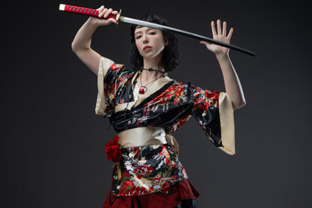 Beautiful model combines traditional Asian aesthetics with cyberpunk fashion in short kimono outfit, wielding katana against plain gray backdrop.の写真素材