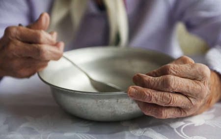 View from above.The hands of an old grandmother of 90 years are holding an empty aluminum bowl and spoon, poverty and poverty, the hunger of the older generation.の写真素材