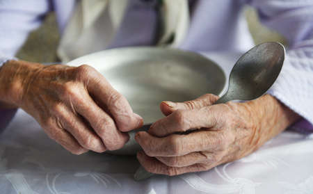 View from above.The hands of an old grandmother of 90 years are holding an empty aluminum bowl and spoon, poverty and poverty, the hunger of the older generation.の写真素材