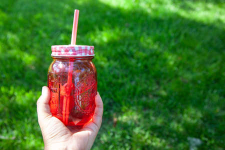 hand holding a red glass jar for cocktails with water or other drink on the background of the lawn.
