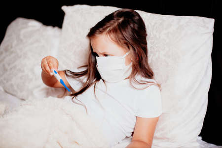 a child in a mask and a white t-shirt on the bed looks at an electronic thermometer, covid-19 or other diseaseの写真素材